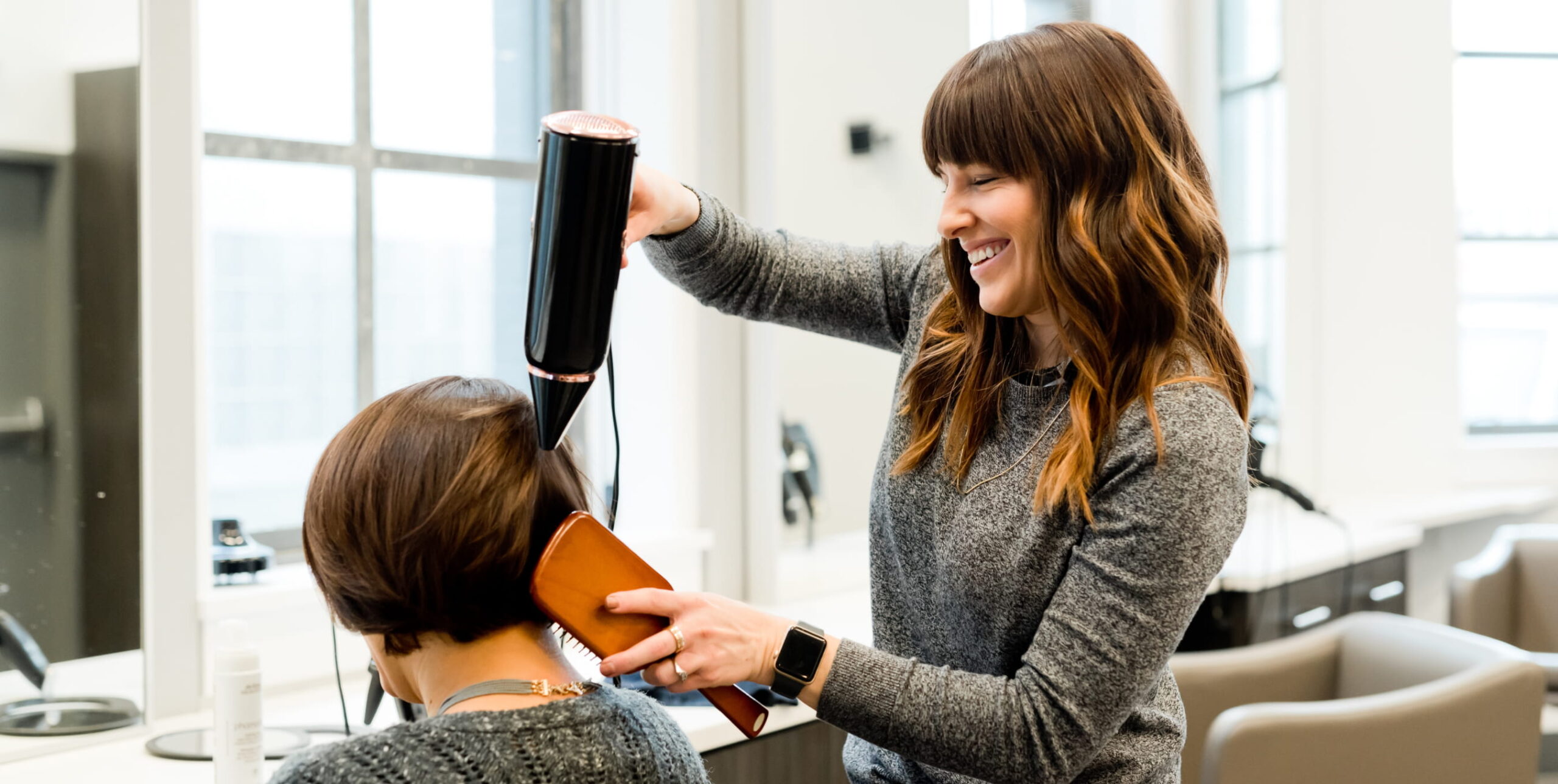 Stylist blow-drying the hair of a woman seated in front of her.